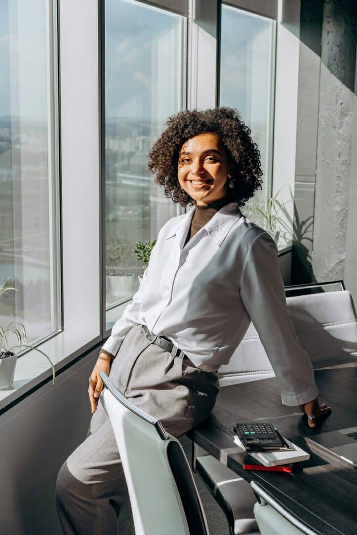 Keep your company's cashflow strong 5 A woman with curly hair, wearing a white blazer and gray pants, smiles while leaning on a desk in a bright office with large windows. Sunlight streams in, illuminating her and the room. A calculator, notebook, pen, and potted plants are on the desk.